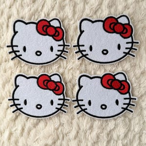 May include: Four white Hello Kitty patches with black outlines, whiskers, and eyes, each with a red bow. The patches are on a textured, off-white surface, ideal for fabric decoration.