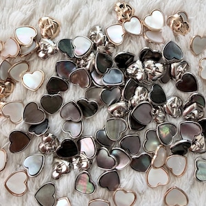 Può includere: Un assortimento di orecchini a forma di cuore in vari colori, tra cui bianco, nero e iridescente, con dettagli in metallo dorato e argentato. Gli orecchini sono sparsi su una superficie bianca e strutturata.