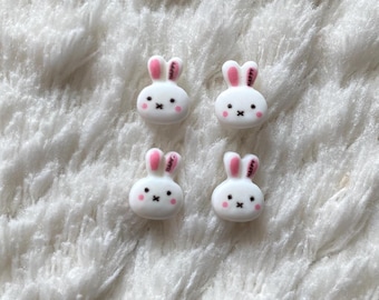 May include: Four white bunny-shaped buttons with pink ears and black eyes. The buttons are on a white fluffy surface.