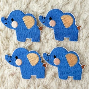 May include: Four blue elephant patches with tan ears and pink cheeks are arranged on a white, textured surface. Each patch has a stitched outline and a small, brown tail. The patches are likely for sewing or crafting.
