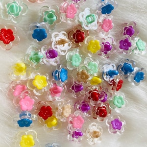 Può includere: Una collezione di bottoni di plastica a forma di fiore di vari colori. I bottoni sono trasparenti e hanno una varietà di colori, tra cui rosso, blu, verde, giallo, viola e rosa.