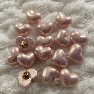 May include: A collection of 14 pink heart-shaped buttons with a pearlescent finish. The buttons have a gold-toned shank on the back.