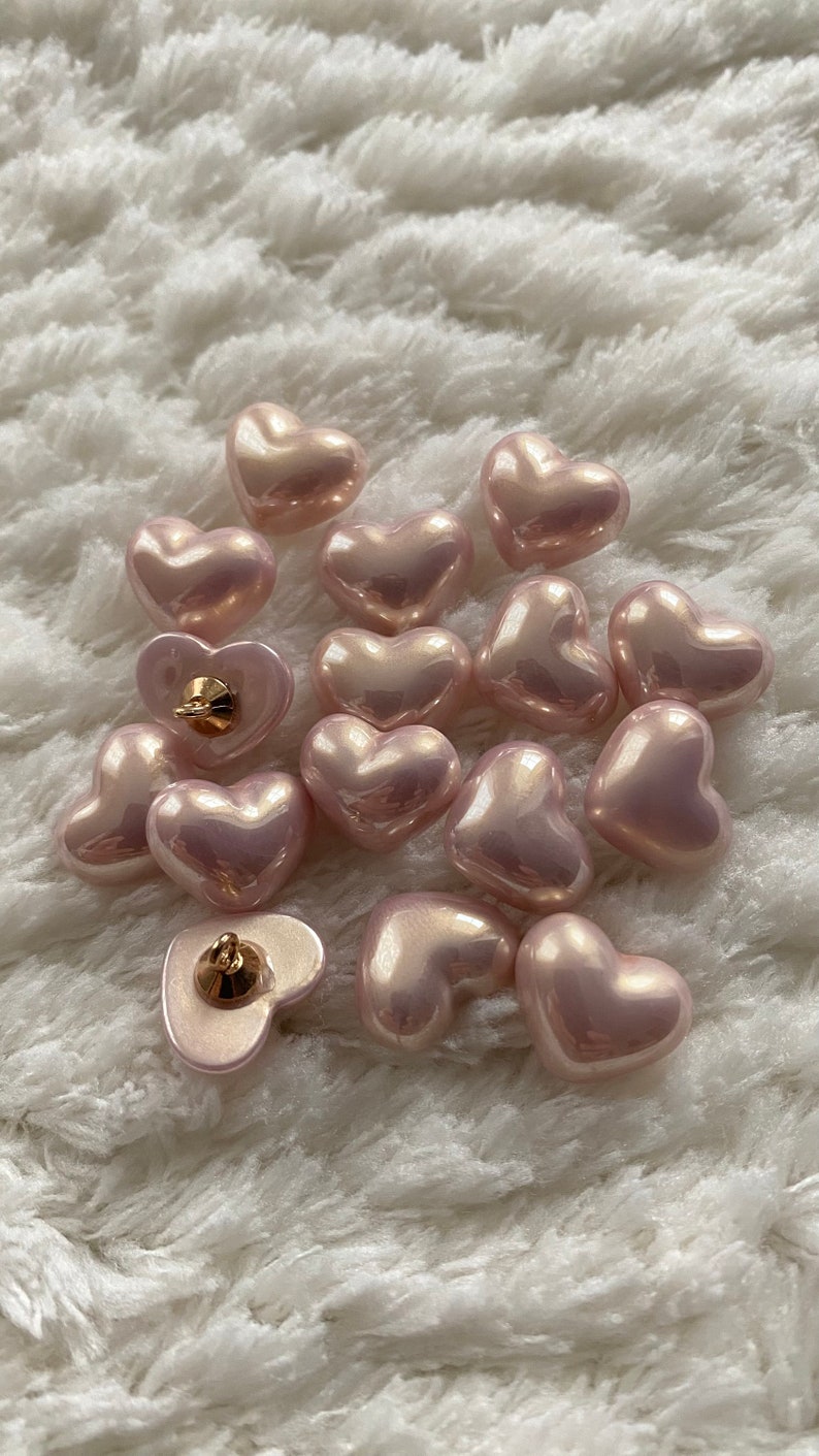 May include: A collection of 14 pink heart-shaped buttons with a pearlescent finish. The buttons have a gold-toned shank on the back.