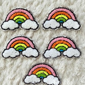 Peut inclure: Cinq écussons arc-en-ciel brodés avec des bordures à paillettes argentées. Chaque écusson présente un motif en arc rose, orange, jaune, vert et bleu reposant sur deux formes blanches texturées rappelant des nuages à la base.