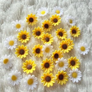May include: A selection of small, decorative flower embellishments. The assortment includes yellow sunflowers with brown centres and white daisies with yellow centres. The flowers are scattered on a soft, white, textured surface.