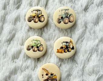 May include: Five round, light-colored buttons with images of construction vehicles. The buttons feature tractors, a bulldozer, and a crane. The vehicles are in shades of yellow, green, and blue. The buttons are on a white, fluffy surface.