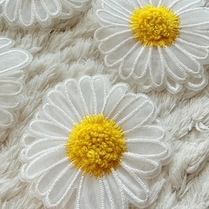 May include: Close-up of several white daisy flower appliques with yellow centers. The petals are outlined with white thread, and the centers have a textured, fuzzy appearance. These floral appliques are perfect for adding a touch of spring to crafts and clothing.