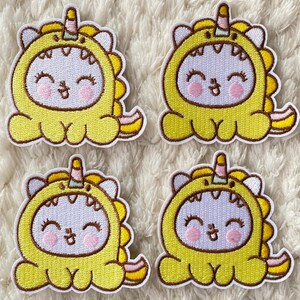 May include: Four yellow embroidered iron-on patches of a cartoon unicorn wearing a dinosaur costume. The unicorn has a pink and white horn, pink cheeks, and a smiling face.