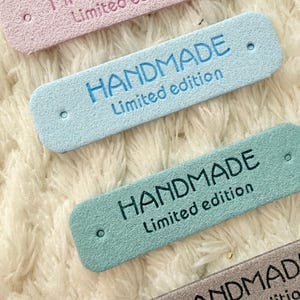 Puede incluir: Primer plano de cuatro etiquetas rectangulares en colores pastel: rosa, azul, verde y gris. Cada etiqueta dice "HANDMADE Limited edition" en un color contrastante. Las etiquetas están sobre una superficie blanca texturizada.