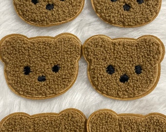 May include: Six brown teddy bear face patches with black embroidered eyes and noses. The patches have a textured, fuzzy appearance and are outlined with a gold-colored border. These patches are ideal for crafting and adding to clothing.
