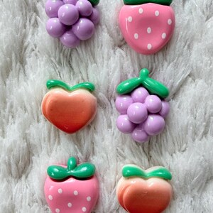 Puede incluir: Una colección de adornos con forma de fruta de colores. La imagen presenta seis piezas: uvas, fresas y melocotones. Las uvas son moradas, las fresas son rosas con puntos blancos y los melocotones son naranjas y rojos.