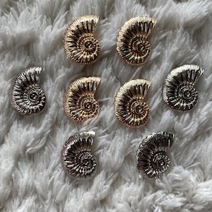 Può includere: Primo piano di otto ciondoli metallici a forma di ammonite. I ciondoli sono in tonalità oro e argento, con dettagliati motivi a spirale. Sono disposti su una superficie bianca e strutturata, creando un contrasto.