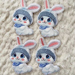 Op de afbeelding: Vier geborduurde patches met een cartoonkonijn met een blauwe gebreide muts en sjaal. De konijnen hebben witte lichamen, roze oren en grote blauwe ogen. De patches liggen op een witte, getextureerde achtergrond.