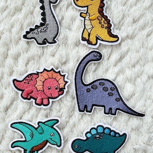 Op de afbeelding: Geborduurde dinosaurussen patches in diverse kleuren, waaronder grijs, geel, roze, paars, turquoise en blauw. Elke patch heeft een ander dinosaurusontwerp met een witte rand. Ideaal voor het toevoegen aan kleding of accessoires.