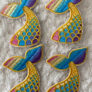 May include: Four colorful mermaid tail patches with a rainbow gradient and yellow scales. The patches are embroidered with a white outline.