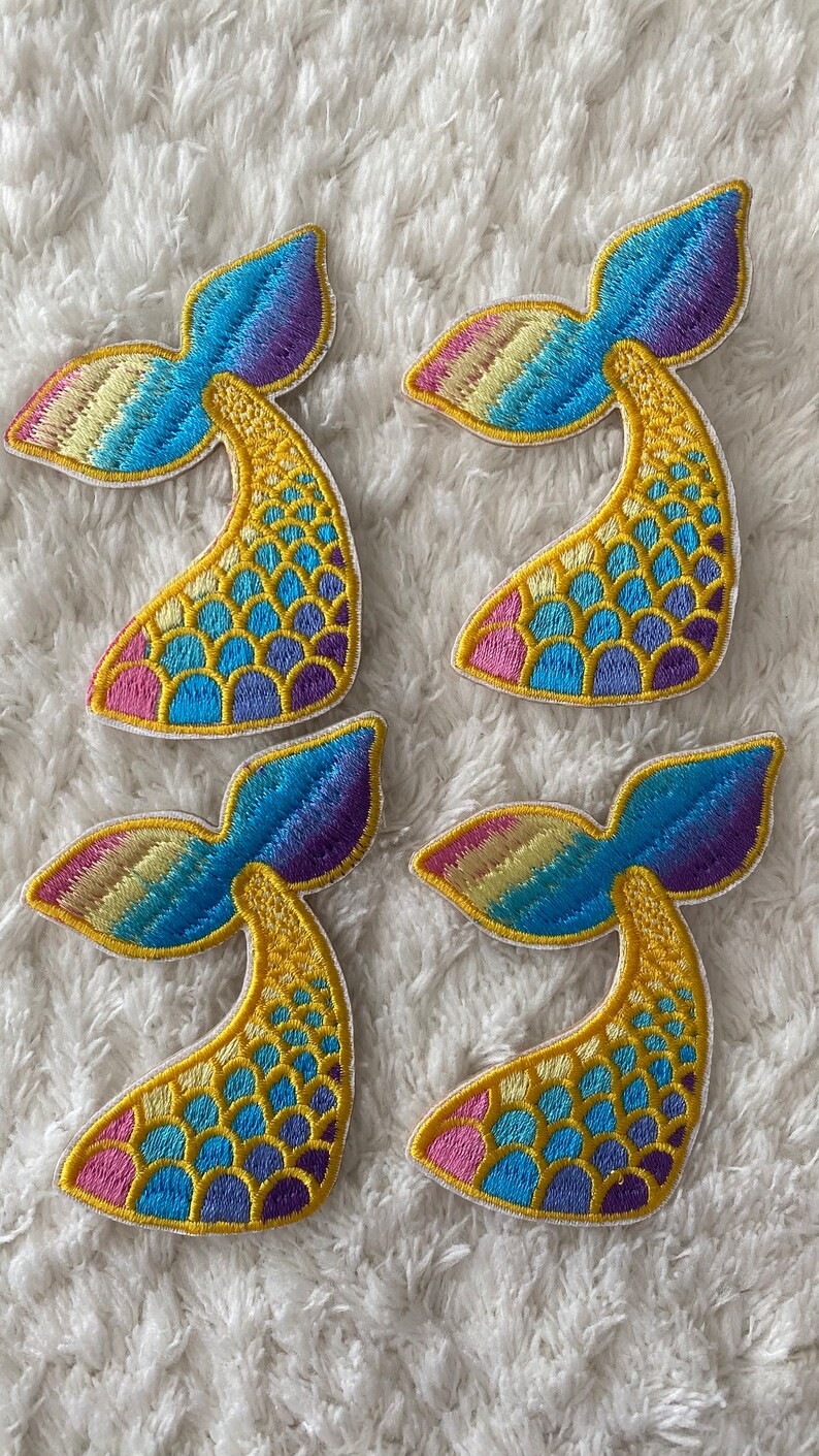 May include: Four colorful mermaid tail patches with a rainbow gradient and yellow scales. The patches are embroidered with a white outline.