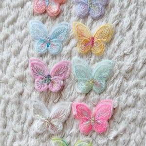 Op de afbeelding: Een verzameling van negen geborduurde vlinderpatches in diverse pastel- en felle kleuren, waaronder perzik, lavendel, lichtblauw, geel, roze, mintgroen, wit en neonroze, elk met gedetailleerd stikwerk en geschulpte randen aan de vleugels.