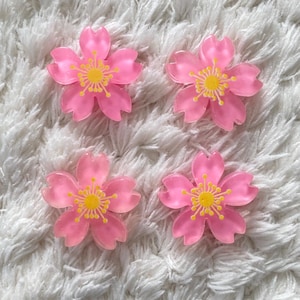 May include: Four pink flower-shaped embellishments with yellow centers are arranged on a white, textured surface. The flowers have five petals each and a translucent appearance. These decorative elements could be used for crafts or embellishments.