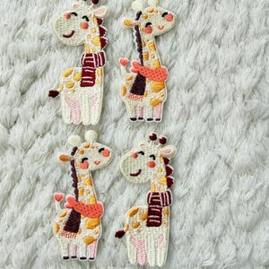 Peut inclure: Quatre écussons brodés de girafes. Chaque girafe a un corps crème avec des taches marron, une écharpe bordeaux et un visage souriant. Les écussons sont sur un fond blanc texturé.