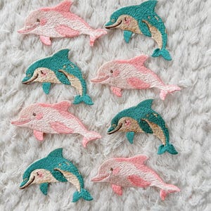 May include: Embroidered dolphin patches in pink and teal. The patches feature a smiling dolphin design with detailed stitching. The dolphins are arranged on a white, textured surface, suitable for sewing onto clothing or accessories.