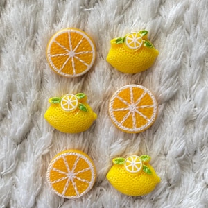 以下が含まれることがあります： 黄色いレモンとオレンジのスライス型の装飾品の詰め合わせ。レモン型のパーツには、小さな白いレモンスライスと緑の葉が付いています。オレンジとレモンのスライスは、白いテクスチャード加工の背景にあります。