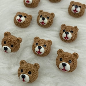 Pode incluir: Dez botões com a forma de cabeça de urso marrom com olhos brancos e boca vermelha. Os botões estão sobre uma superfície branca e felpuda.