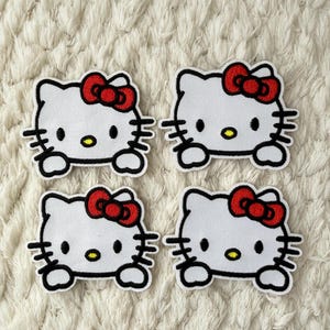 May include: Four white embroidered patches featuring the iconic Hello Kitty design. Each patch has a red bow, black whiskers, and a yellow nose. The patches are set against a textured, off-white background.