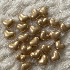May include: A collection of gold heart-shaped buttons. The buttons are smooth and shiny, and they are arranged in a random pattern on a white fuzzy surface.