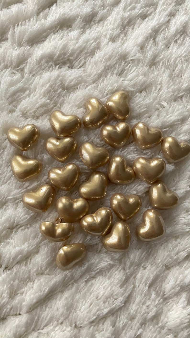 May include: A collection of gold heart-shaped buttons. The buttons are smooth and shiny, and they are arranged in a random pattern on a white fuzzy surface.