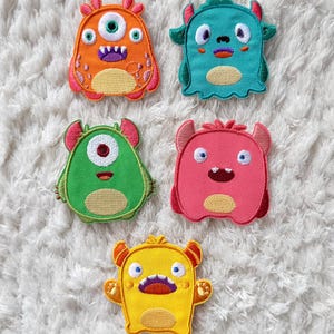 May include: Five colorful monster-shaped embroidered patches. The patches are orange, teal, green, pink, and yellow. Each monster has a unique design with horns, eyes, and mouths. The patches are on a white, fluffy surface.