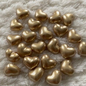May include: A group of gold heart-shaped beads on a white fluffy surface.