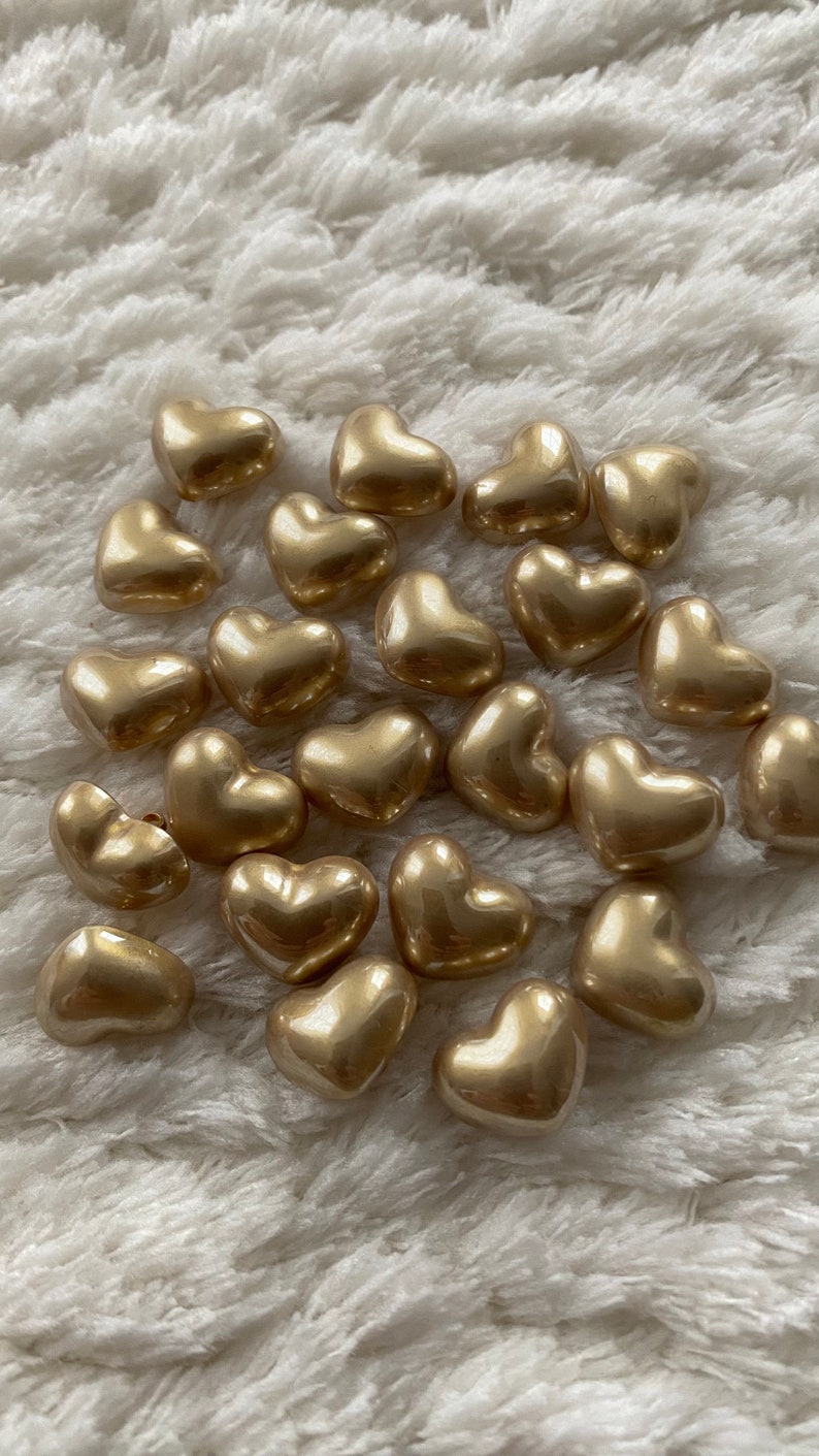 May include: A group of gold heart-shaped beads on a white fluffy surface.
