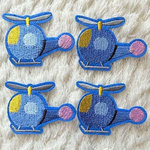 May include: Four embroidered patches in the shape of a blue helicopter with yellow and pink accents. The patches are on a white, textured surface. The helicopter patches have a propeller, a tail, and a small window.