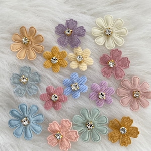 Könnte beinhalten: Sortiment von 14 Stoffblumen-Verzierungen mit Strasssteinen in verschiedenen Pastellfarben, darunter hellblau, rosa, Lavendel, gelb und grün. Die Blumen sind auf einer weißen, flauschigen Oberfläche angeordnet.