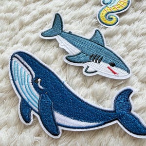 以下が含まれることがあります： 海の生き物をあしらった刺繍パッチ：青いクジラ、サメ、タツノオトシゴ。クジラは濃い青色に白いアクセント、サメは水色に白いお腹と赤い口、タツノオトシゴは黄色と青色です。