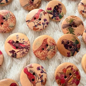 Könnte beinhalten: Eine Sammlung runder Holzknöpfe mit Vogelillustrationen. Jeder Knopf zeigt ein anderes Vogeldesign in Rot-, Braun- und Grüntönen mit floralen Akzenten. Die Knöpfe haben zwei Löcher zum Annähen.