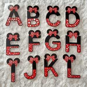May include: A set of 12 iron-on patches featuring the letters A through L in a black and red polka dot pattern with a red bow on top. The letters are on a clear background.