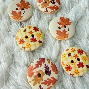 Könnte beinhalten: Fünf runde Holzknöpfe mit jeweils zwei Löchern. Drei Knöpfe haben einen cremefarbenen Hintergrund mit verschiedenen orangefarbenen, roten und violetten Herbstblattmotiven. Zwei Knöpfe zeigen ein gelb-weißes Karomuster mit kleinen roten und orangen Blättern.