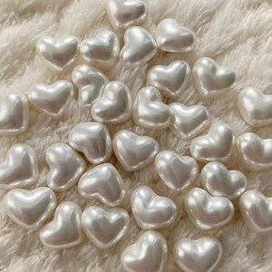 May include: A collection of white pearl heart-shaped beads on a white fuzzy surface.