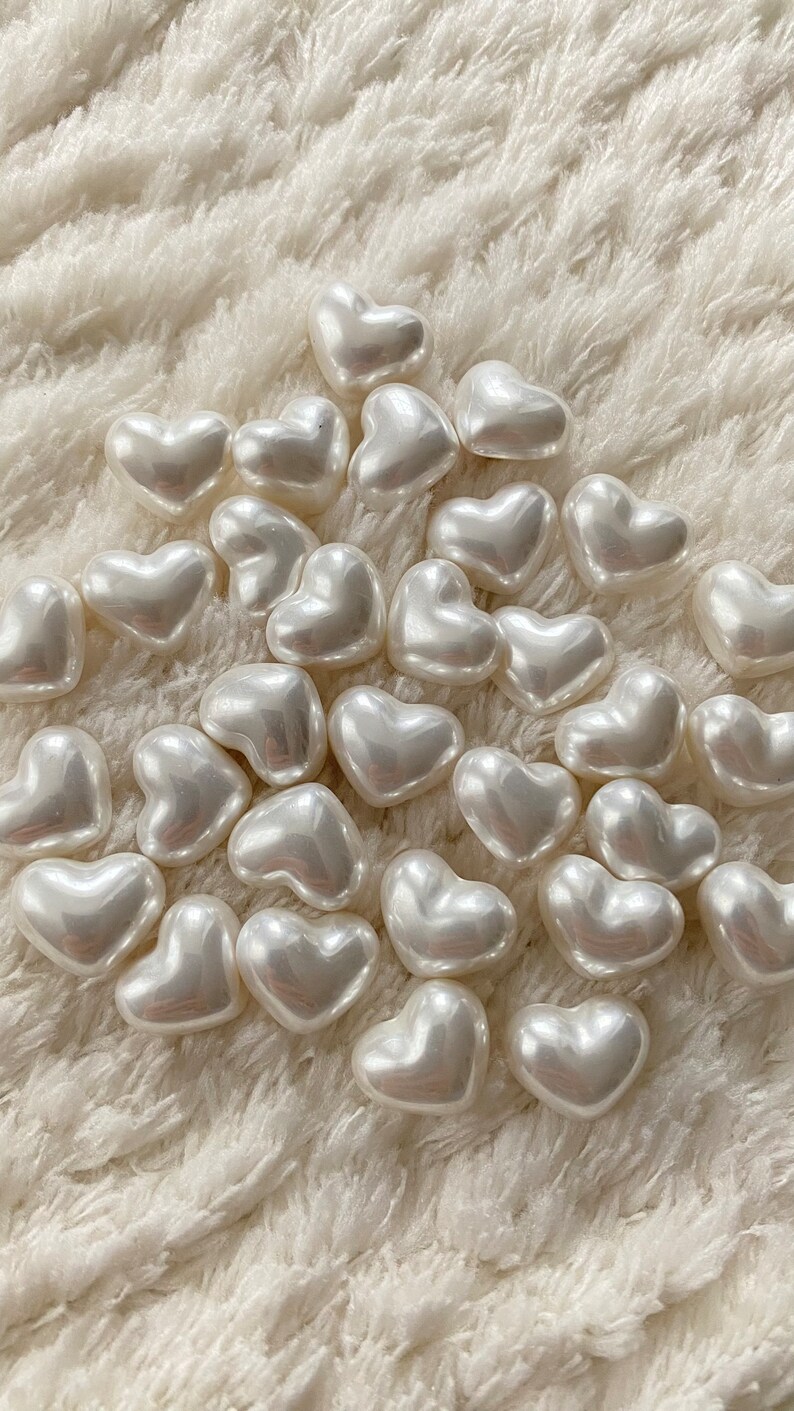 May include: A collection of white pearl heart-shaped beads on a white fuzzy surface.