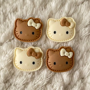 Può includere: Quattro ciondoli in resina in stile Hello Kitty. Due sono marroni con fiocchi bianchi e due sono bianchi con fiocchi marroni.