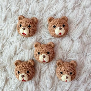 Pode incluir: Cinco enfeites de rosto de urso de pelúcia marrons com olhos pretos, boca vermelha e nariz bege. Os rostos dos ursos estão dispostos sobre uma superfície branca e texturizada.