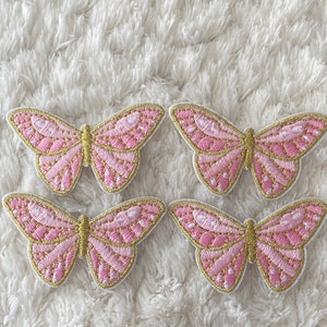 May include: Four pink and gold butterfly iron-on patches. The butterflies have a detailed design with pink and gold embroidery.