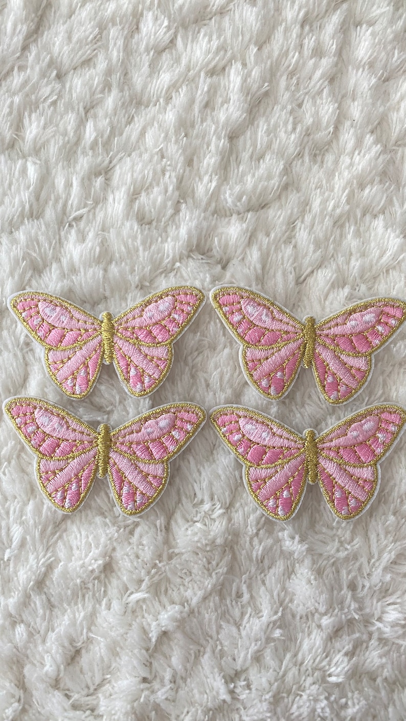 May include: Four pink and gold butterfly iron-on patches. The butterflies have a detailed design with pink and gold embroidery.