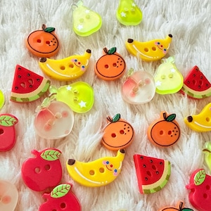 Puede incluir: Una colección de pequeños botones coloridos con forma de fruta, que incluye plátanos amarillos con caras, rodajas de cítricos naranjas, formas de manzana roja, cuñas de sandía y formas de pera o melocotón translúcidas, cada uno con dos agujeros.