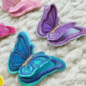 May include: Embroidered butterfly patches in shades of pink, purple, and blue. The patches have detailed stitching and are arranged on a white, textured surface. These patches are ideal for adding a decorative touch to clothing or accessories.