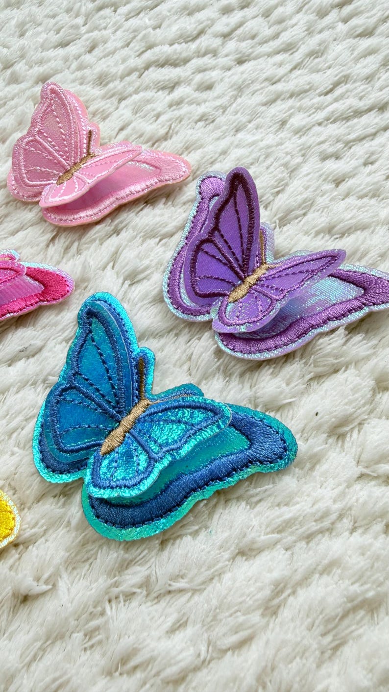 May include: Embroidered butterfly patches in shades of pink, purple, and blue. The patches have detailed stitching and are arranged on a white, textured surface. These patches are ideal for adding a decorative touch to clothing or accessories.