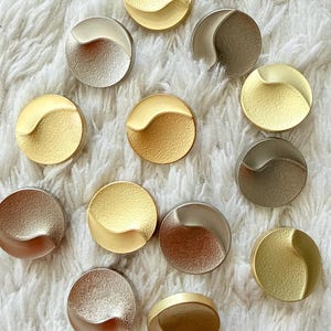 Peut inclure: Une collection de boutons circulaires aux finitions métalliques variées, notamment or, argent et bronze. Chaque bouton présente une crête incurvée et surélevée distincte qui divise la surface en deux sections, l'une lisse et l'autre texturée.