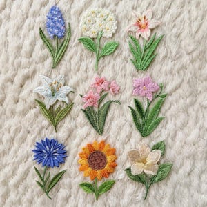 Könnte beinhalten: Eine Sammlung von neun gestickten Blumenaufnähern in verschiedenen Farben, darunter Blau, Weiß, Rosa, Gelb und Orange. Jeder Aufnäher weist detaillierte Stickereien und grüne Blätter auf und zeigt verschiedene Blumenarten wie Lilien, Sonnenblumen und Hortensien. Die Aufnäher sind auf einer strukturierten, cremefarbenen Oberfläche angeordnet.