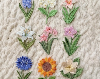 May include: A collection of nine embroidered flower patches in various colors, including blue, white, pink, yellow, and orange. Each patch features detailed stitching and green leaves, showcasing different flower types like lilies, sunflowers, and hydrangeas. The patches are arranged on a textured, off-white surface.